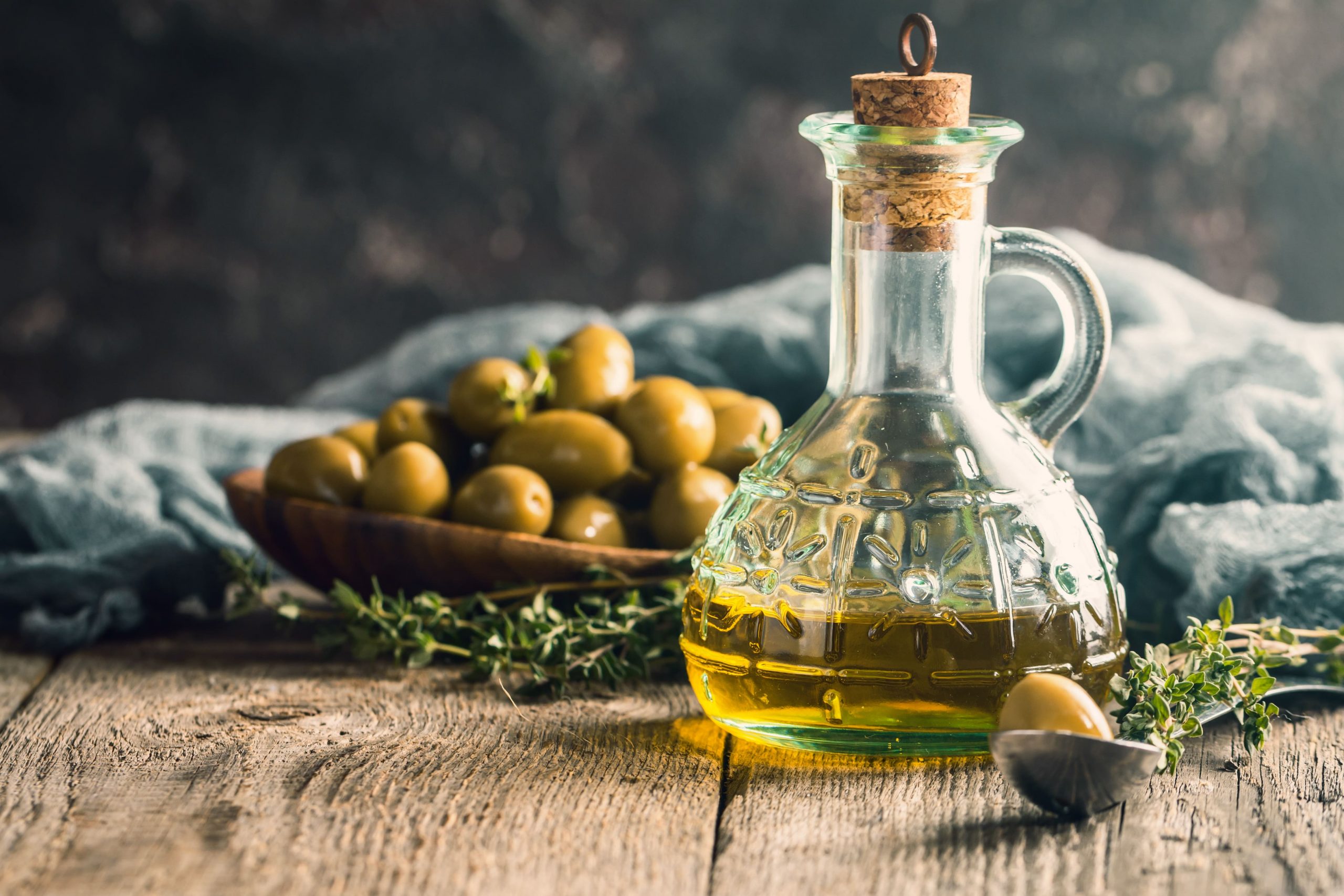 frasco de aceite de oliva sobre una mesa junto con una cuchara y un recipiente de olivas verdes