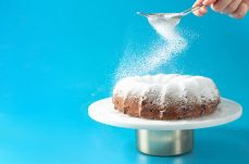 Woman's hand sprinkling icing sugar over fresh home made bundt cake. Powder sugar falls on fresh perfect bunt cake over blue background. Copy space for text. Ideas and recipes for breakfast or dessert